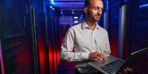 Caucasian programmer is looking to the screen of laptop while standing near the server rack